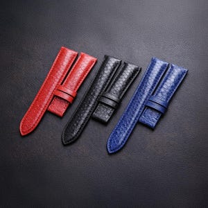 Könnte beinhalten: Drei Uhrenarmbänder in Rot, Schwarz und Blau. Jedes Armband hat ein strukturiertes, reptilienartiges Muster und eine passende Schnalle. Die Armbänder werden auf einer dunklen, strukturierten Oberfläche präsentiert, die ihre Farben und ihr Design hervorhebt.