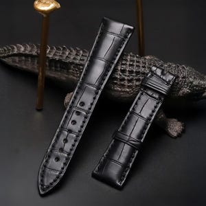 May include: Two-piece black watch strap with a crocodile skin texture. The strap has a buckle and several holes for adjustment. The stitching is visible along the edges. The background includes a decorative alligator sculpture and gold-colored accents.