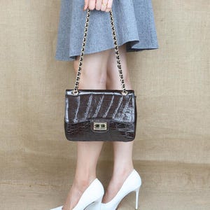 May include: A dark brown, rectangular handbag with a crocodile skin texture. The bag has a gold chain strap and a gold clasp. The person is wearing a gray skirt and white high heels.