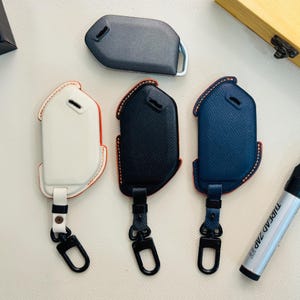 Op de afbeelding: Drie gestructureerde lederen autosleutelhoezen in wit, zwart en marineblauw, elk met contraststiksels, een bijpassende lederen riem en een zwarte metalen draaibare karabijnhaak. Een grijze plastic sleutelbehuizing ligt daarboven.
