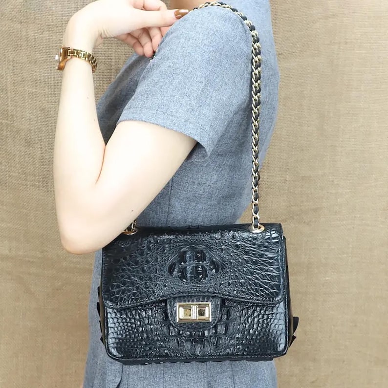 May include: Black leather crossbody bag with a crocodile skin texture. The bag features a gold-tone clasp and a chain strap with black leather accents. The bag is rectangular and has a flap closure.