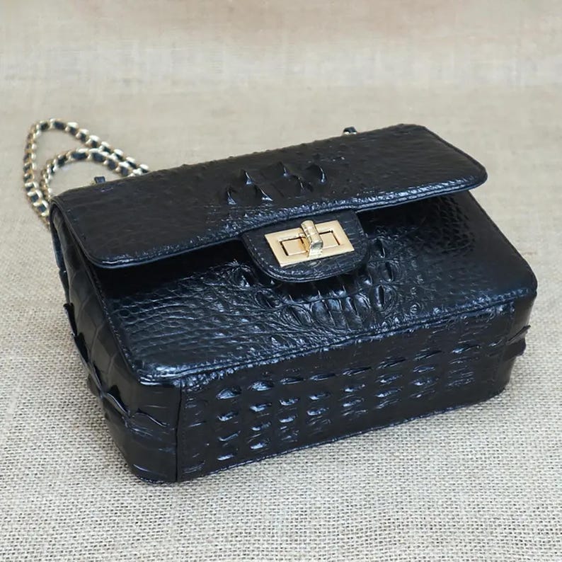 May include: Black leather handbag with a textured crocodile pattern. The bag features a gold-tone clasp and chain strap. The rectangular bag has a flap closure and is designed for carrying essentials.