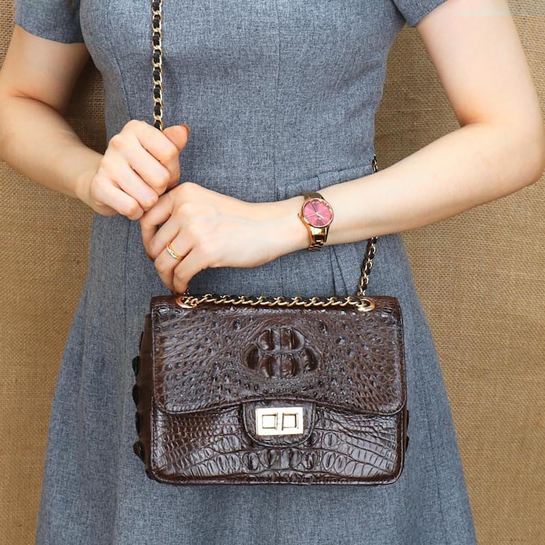 May include: A brown leather crossbody bag with a crocodile skin texture. The bag has a gold chain strap and a gold clasp closure. The watch on the wrist has a pink face and a gold band.