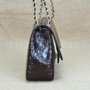 May include: A brown leather handbag with a textured, crocodile-like pattern. The bag features a gold chain strap with black accents and a flap closure. The bag is rectangular and has a classic design.