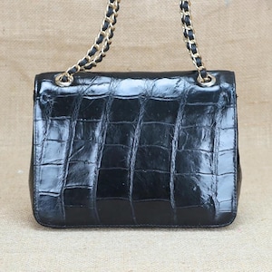 May include: A black, rectangular handbag with a textured, crocodile-like pattern. The bag features a gold chain strap with black leather accents. The bag is glossy and appears to be made of leather.