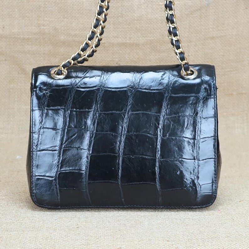 May include: A black, rectangular handbag with a textured, crocodile-like pattern. The bag features a gold chain strap with black leather accents. The bag is glossy and appears to be made of leather.