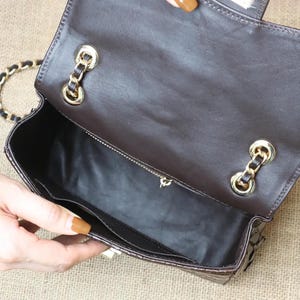 May include: Open view of a brown leather handbag with a gold chain strap. The interior reveals a black lining and a zippered pocket. Gold-toned hardware accents the bag's closure and strap attachments.