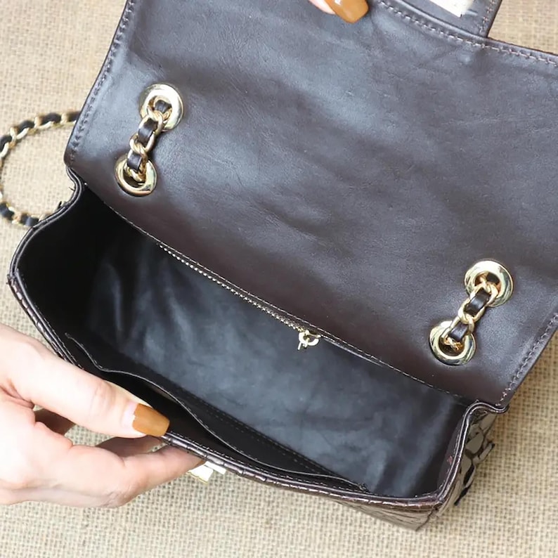 May include: Open view of a brown leather handbag with a gold chain strap. The interior reveals a black lining and a zippered pocket. Gold-toned hardware accents the bag's closure and strap attachments.