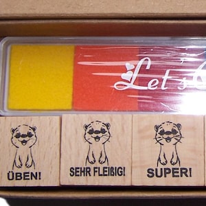 Könnte beinhalten: Ein Set mit farbigen Stempelkissen und Holzstempeln in einer Schachtel. Die Stempelkissen sind gelb, orange, rot, violett, blau und grün. Die Holzstempel zeigen einen Otter und deutschen Text.