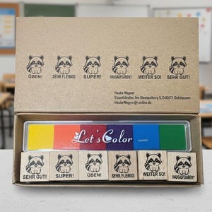 Könnte beinhalten: Ein Bastelset mit einem mehrfarbigen Stempelkissen und Holzstempeln. Die Stempel zeigen Waschbär-Illustrationen und deutsche Sätze. Die Box ist geöffnet und zeigt die Stempel und das Stempelkissen mit dem Text "Let's Color".