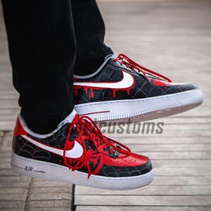 Puede incluir: Zapatillas personalizadas negras y rojas con detalles blancos. Los zapatos presentan un dise&ntilde;o de telara&ntilde;a y detalles de goteo de pintura roja. Las suelas blancas tienen la palabra "AIR" impresa en el lateral.