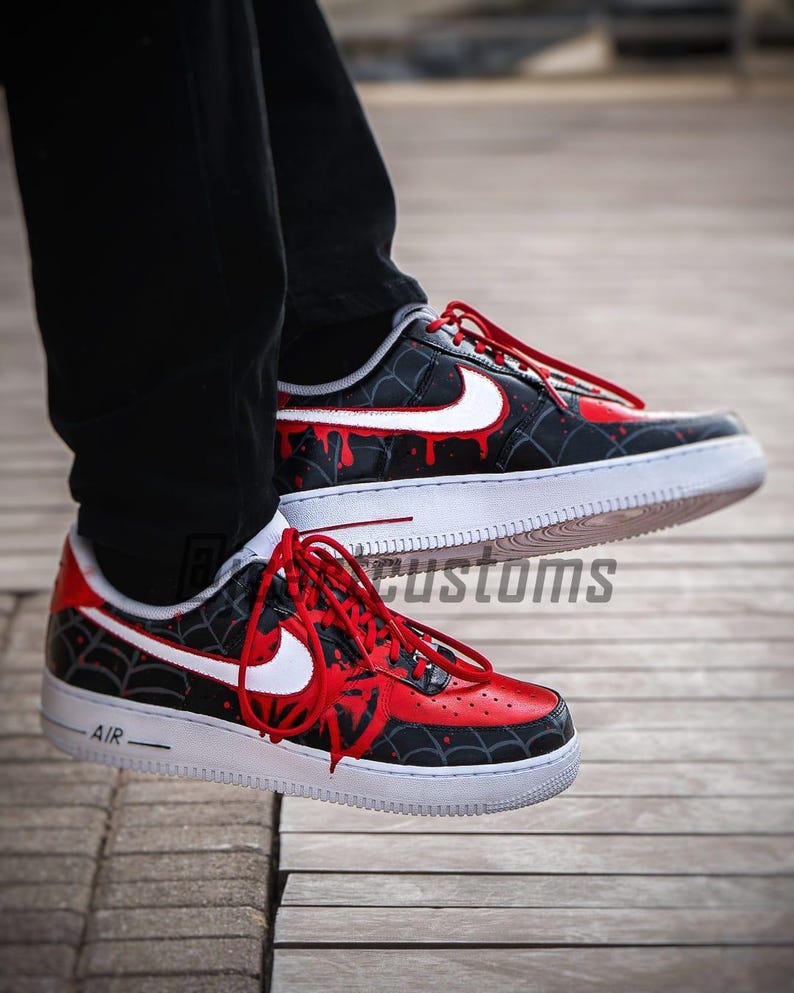 Puede incluir: Zapatillas personalizadas negras y rojas con detalles blancos. Los zapatos presentan un dise&ntilde;o de telara&ntilde;a y detalles de goteo de pintura roja. Las suelas blancas tienen la palabra "AIR" impresa en el lateral.