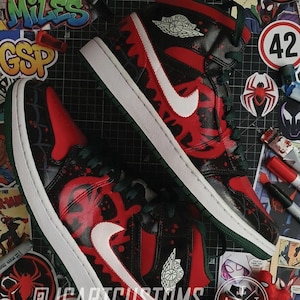 May include: Pair of custom Air Jordan 1 sneakers in a Spider-Verse theme. The shoes feature a red and black design with white accents, a white Nike swoosh, and green laces. The soles are white with green trim. The image includes stickers and markers.