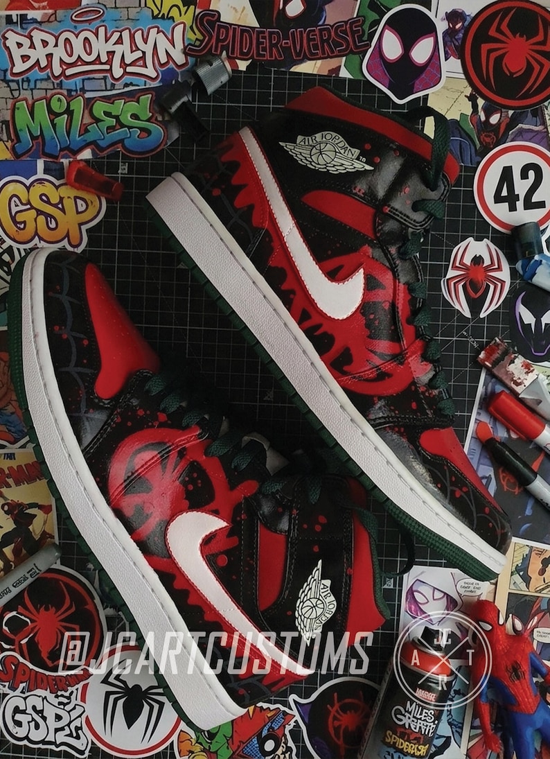 May include: Pair of custom Air Jordan 1 sneakers in a Spider-Verse theme. The shoes feature a red and black design with white accents, a white Nike swoosh, and green laces. The soles are white with green trim. The image includes stickers and markers.