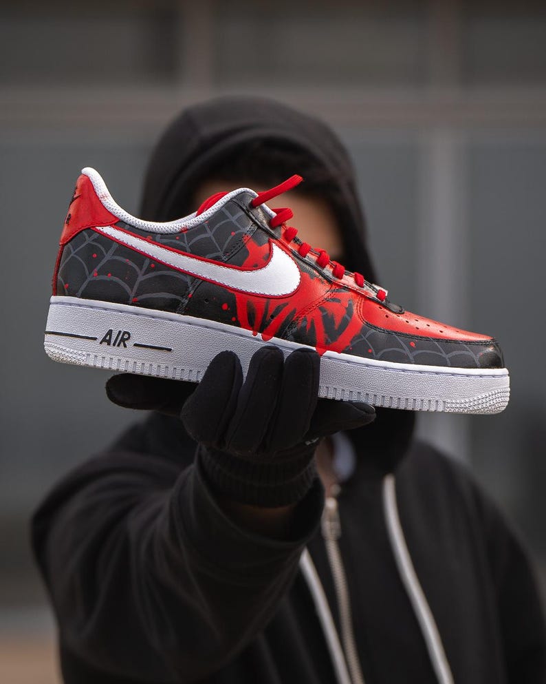Puede incluir: Una zapatilla pintada a medida con un dise&ntilde;o negro y rojo con un patr&oacute;n de telara&ntilde;a. La zapatilla tiene una suela blanca y un logotipo de Nike blanco. La palabra "AIR" est&aacute; impresa en el lateral. Los cordones rojos completan el look.