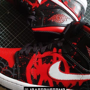 May include: Close-up of custom black and red sneakers with white accents. The shoes feature a red splatter design and a white Nike swoosh. The text "@JCARTCUSTOMS" is at the bottom.
