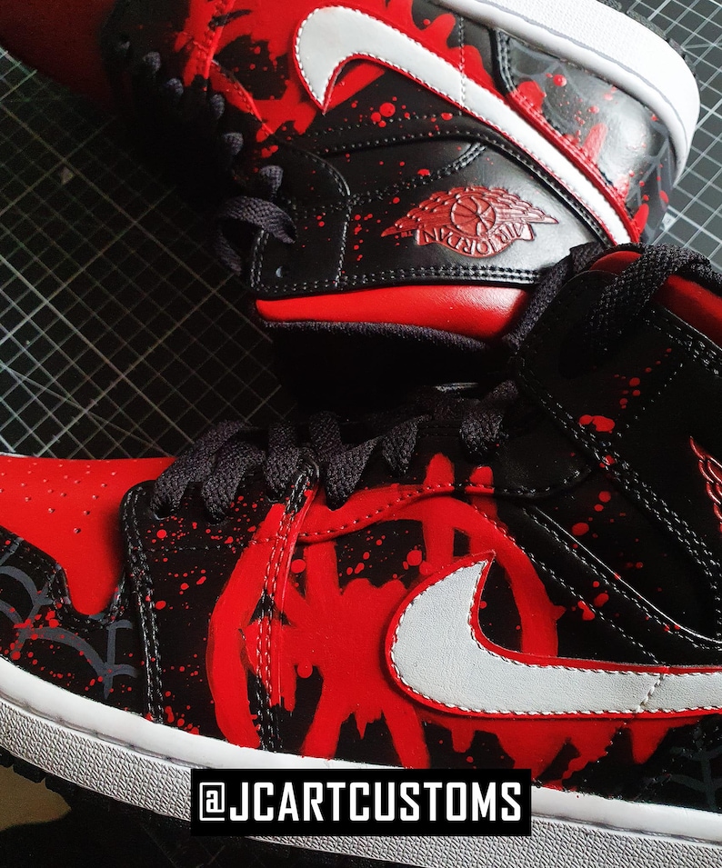 May include: Close-up of custom black and red sneakers with white accents. The shoes feature a red splatter design and a white Nike swoosh. The text "@JCARTCUSTOMS" is at the bottom.
