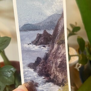 Puede incluir: Pequeña pintura rectangular de un paisaje costero. La obra representa un acantilado rocoso que se encuentra con el océano bajo un cielo nublado. La pintura utiliza una paleta de azules, grises y marrones para crear una escena serena.