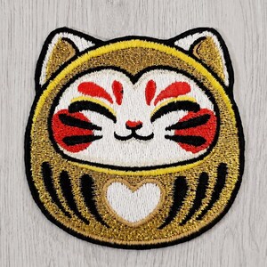 May include: An embroidered patch shaped like a round cat face with a Daruma-inspired design. The cat features gold metallic thread, a white face with red and black markings, pointed ears, and a white heart shape on its chest, all outlined in black.