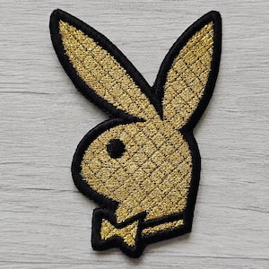 Könnte beinhalten: Ein gestickter Aufnäher mit der ikonischen Hasenkopf-Silhouette aus metallischem Goldfaden mit schwarzem Rand. Das Design umfasst die langen Ohren des Hasen, ein kreisförmiges Auge und eine kleine Fliege an der Basis, alles in strukturierter Stickerei.