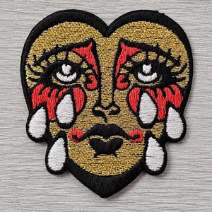 May include: An embroidered patch in the shape of a heart featuring a stylised face with gold skin, black outlines, and red accents around the eyes and mouth. The design includes white teardrops falling from the eyes and cheeks, all bordered in black.