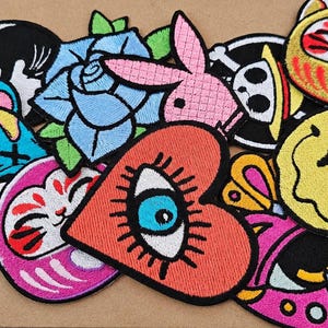 Pode incluir: Uma coleção de emblemas de tecido bordado com diversos designs, incluindo uma rosa azul, um coelho rosa, um coração vermelho com um olho central, um rosto sorridente amarelo, duas figuras felinas, uma caveira e uma forma abstrata preta e branca.