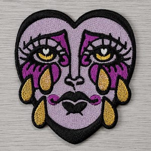 May include: An embroidered patch in the shape of a heart featuring a stylised face with lavender skin, dark purple eyeshadow, and black eyelashes. The face has a black mouth and is shedding gold teardrops from both eyes, outlined in black thread.
