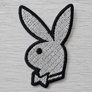 Könnte beinhalten: Ein silber-schwarzer Stickaufnäher, der die ikonische Silhouette eines Hasenkopfes mit langen Ohren und einer Fliege zeigt. Das Design ist in strukturiertem, metallisch silbernem Garn gehalten und von einem schwarzen Rand umgeben.