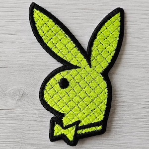 Könnte beinhalten: Ein gestickter Aufnäher, der die ikonische Hasenkopf-Silhouette in leuchtendem Neongrün mit einer gesteppten Rautenstruktur zeigt. Das Design ist mit einem dicken schwarzen Garnrand umrandet und enthält ein kleines schwarzes Auge.