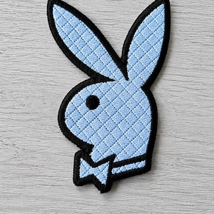 Könnte beinhalten: Ein hellblau bestickter Aufnäher, der die ikonische Hasenkopf-Silhouette mit einer Fliege zeigt. Das Design weist eine gesteppte Rautenstruktur über die gesamte Hasenform auf, die von einem dicken schwarzen Rand umgeben ist und Kontrast bietet.
