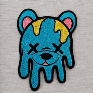 Op de afbeelding: Geborduurde patch met een blauwe beer met gele druppeldetails. De beer heeft zwarte kruizen voor de ogen en een roze tong. De patch heeft een zwarte omtrek en ligt op een lichtgrijze houten ondergrond.