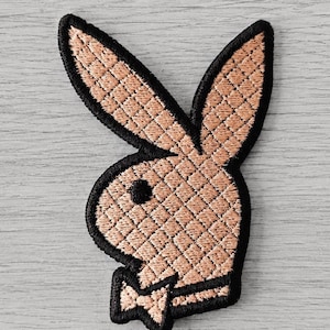 Könnte beinhalten: Ein gestickter Aufnäher mit der Silhouette eines Kaninchenkopfes im Profil, in einem hellen Beigeton mit gestepptem Rautenmuster. Das Design ist mit dickem schwarzem Garn umrandet und zeigt ein schwarzes Auge sowie eine schwarze Fliege am Hals.
