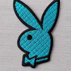 Könnte beinhalten: Ein gestickter Aufnäher mit der ikonischen Hasenkopf-Silhouette aus leuchtend türkisfarbenem Garn mit schwarzem Rand und einer kleinen schwarzen Fliege. Das Hasenmotiv weist ein strukturiertes Kreuzstichmuster über die gesamte Form auf.