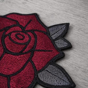 Puede incluir: Parche bordado con una rosa roja detallada con contornos negros y hojas grises. La rosa tiene una apariencia texturizada, ideal para ropa o accesorios.