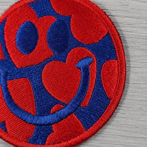 Puede incluir: Parche bordado con diseño rojo y azul. El parche circular tiene un borde rojo. El diseño interior presenta corazones rojos y caras sonrientes azules.