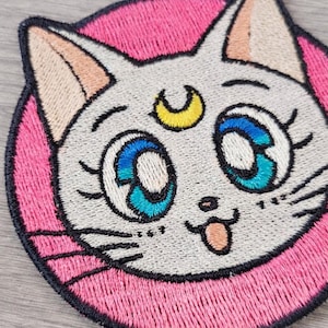 May include: Embroidered patch of a cartoon cat's face, with large blue eyes, a yellow crescent moon, and a pink circular background. The cat has a light grey face, pink ears, and a small pink tongue.