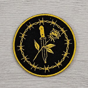 Puede incluir: Parche bordado con fondo negro y borde amarillo. El diseño presenta un cuchillo amarillo que perfora una rosa amarilla, rodeado por un círculo de alambre de púas. Las hojas también están incluidas en el diseño.