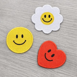 May include: Three embroidered patches with smiling faces. A yellow circle, a white daisy with a yellow center, and a red heart, all with black stitched smiles. These patches are likely for clothing or accessories.
