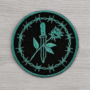 Könnte beinhalten: Gestickter Aufnäher mit einem türkisfarbenen Dolch und einer Rose, umgeben von türkisfarbenem Stacheldraht auf schwarzem Grund. Auf hellem Holzmaserungshintergrund. Durchmesser ca. 7 cm.