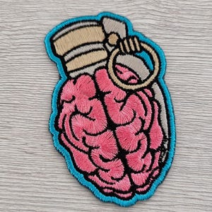 May include: Embroidered patch featuring a pink brain design within a grenade shape. The patch has a light blue border, with tan and gray details. The brain is detailed with black outlines. The patch is likely for clothing or accessories.