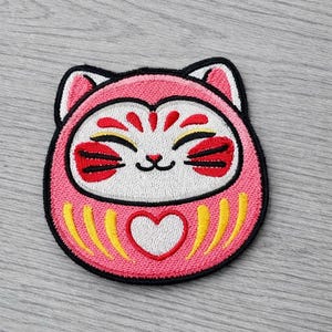 May include: Embroidered patch featuring a pink Daruma doll design. The doll has a white face with a smiling expression, red accents, and a heart shape. The patch is outlined in black.