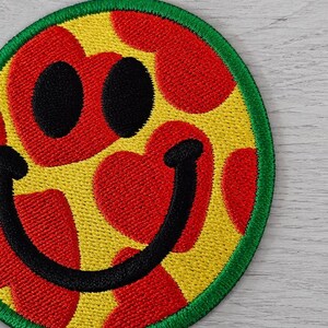 May include: Embroidered patch featuring a smiling face with a pizza design. The patch has a yellow background with red heart-shaped pepperoni, black eyes, and a black curved mouth. A green border surrounds the design.