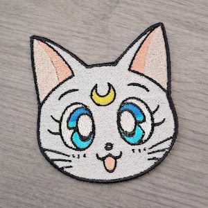 May include: Embroidered patch depicting a cartoon cat's face with large blue eyes, a yellow crescent moon, and a pink tongue. The cat has white fur with black outlines and pink inner ears. Suitable for clothing or accessories.