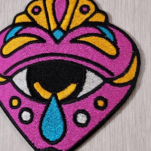 Puede incluir: Parche bordado en forma de corazón, con un ojo estilizado y una lágrima. El diseño incorpora colores rosa, negro, amarillo, blanco y azul. El parche tiene una corona decorativa en la parte superior y pequeños círculos alrededor del ojo.