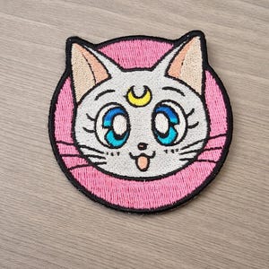Può includere: Toppa ricamata raffigurante un volto di gatto bianco con occhi blu, una luna crescente gialla e un bordo circolare rosa. Il gatto ha una piccola lingua rosa che sporge. La toppa è su una superficie di legno.
