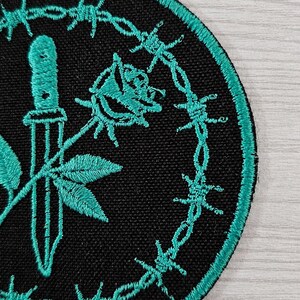 May include: Embroidered patch with a black background and a teal border. The patch features a teal dagger, rose, and leaves, surrounded by a teal barbed wire design. The patch is circular.