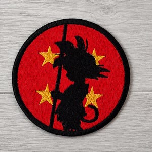 Può includere: Toppa ricamata con una silhouette nera di un personaggio che tiene un bastone su sfondo rosso. Quattro stelle gialle sono disposte attorno al personaggio all'interno di un cerchio bordato di nero.
