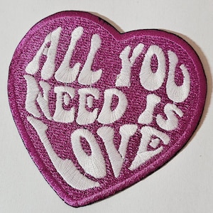 Könnte beinhalten: Ein herzförmiger Aufnäher mit der gestickten Aufschrift „ALL YOU NEED IS LOVE“. Der Aufnäher hat einen leuchtend pinkfarbenen Hintergrund und weiße Buchstaben. Er ist für Kleidung oder Accessoires gedacht.