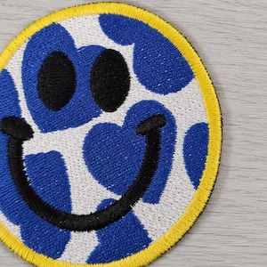 Pode incluir: Patch bordado com um design de rosto sorridente. O rosto tem olhos pretos e um sorriso preto. O fundo é branco com formas de coração azuis. O patch é contornado em amarelo.