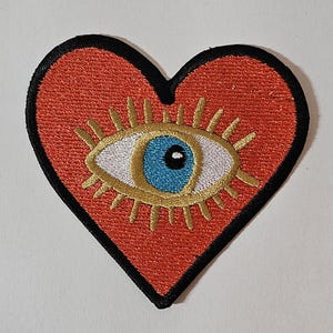 May include: Embroidered heart-shaped patch with an eye design. The heart is red with a black border. The eye has a blue iris, white and gold details. A decorative accessory.